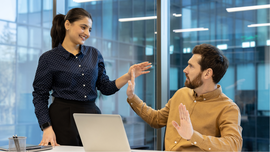 /_cms/webroot/In an office, a standing person leans over a seated person and reaches for them, while the seated person reacts with a raised hand in refusal.